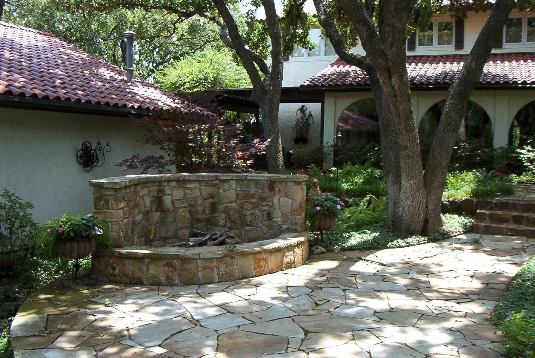 Curved stone fountain and flagstone patio under mature oaks beside a stucco house