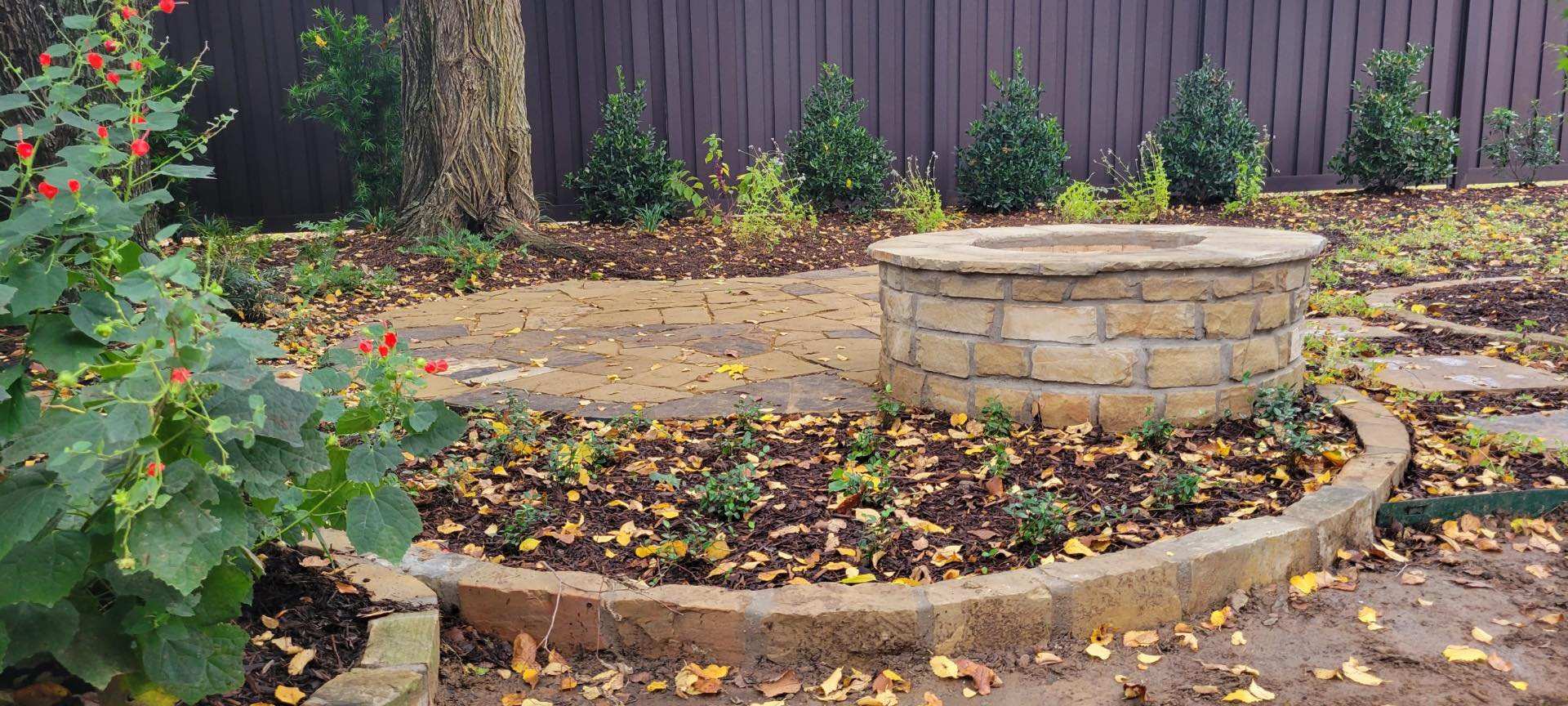 Raised circular stone planter with a young tree in a mulched bed against a dark fence